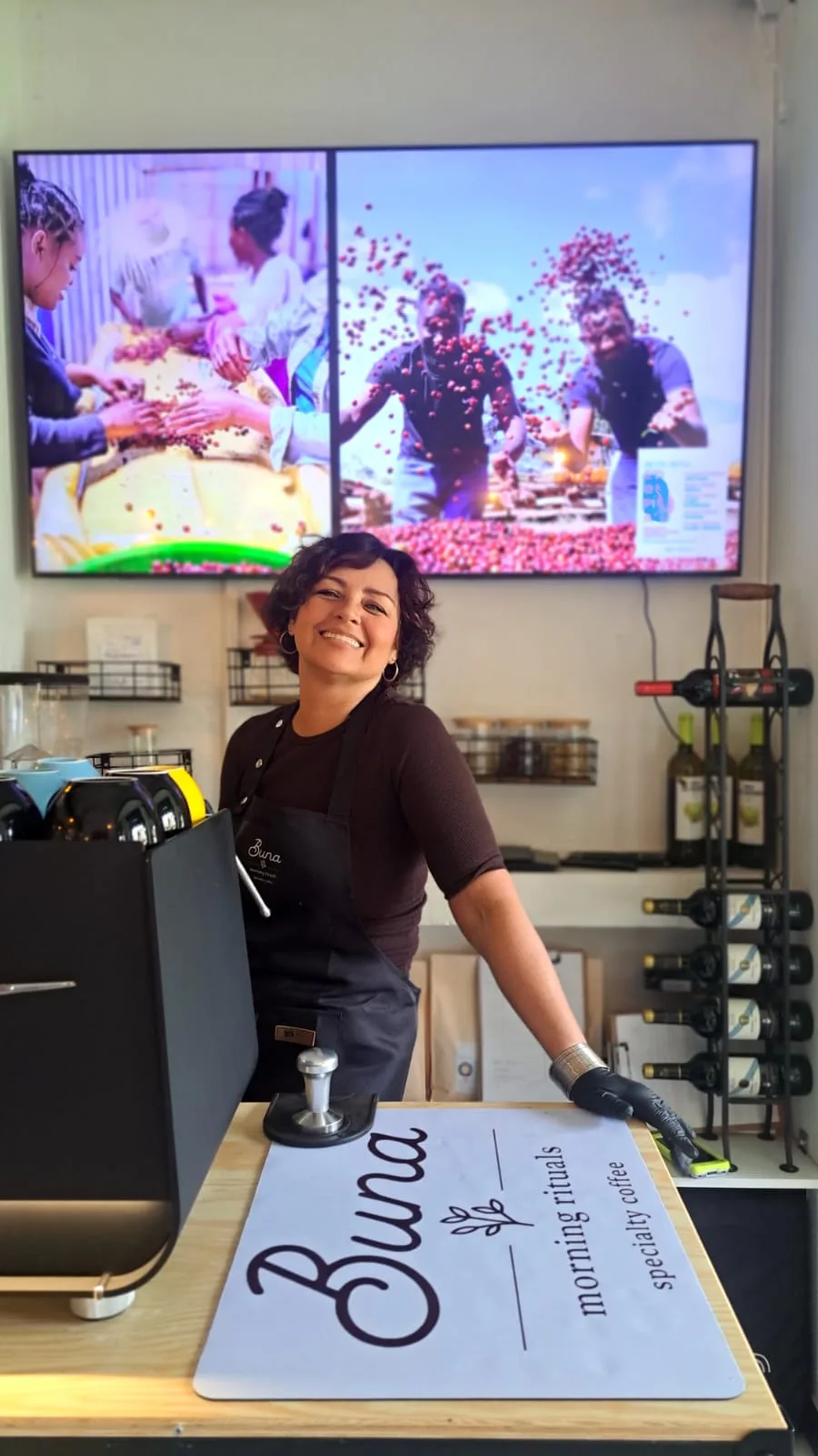 Barista de Buna sonriendo tras la barra, junto a la máquina de espresso y la bandeja con el logotipo de Buna
