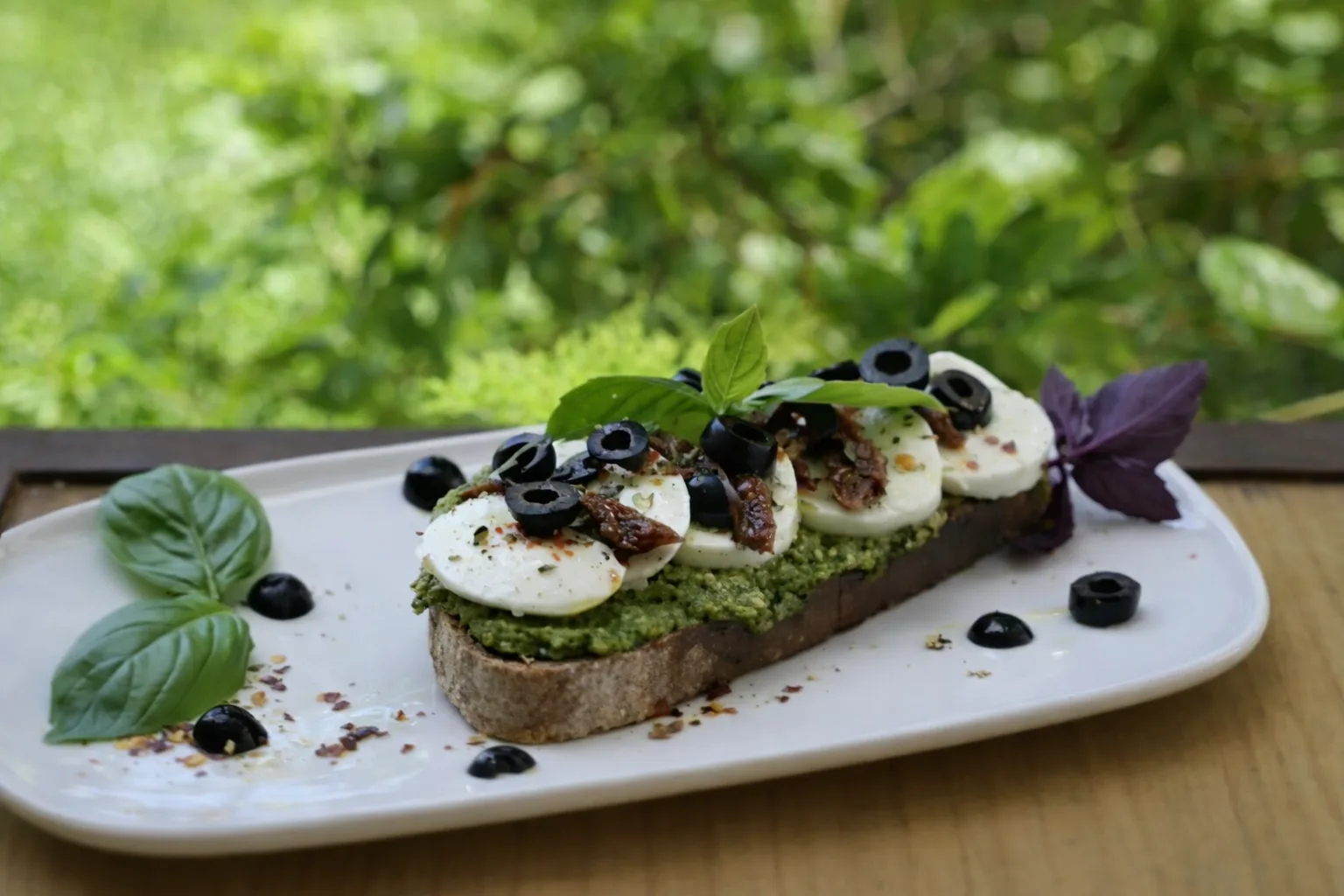 Tosta de pan de masa madre con pesto, mozzarella, tomate seco, aceitunas negras y albahaca, en un plato blanco sobre una mesa de madera del jardín