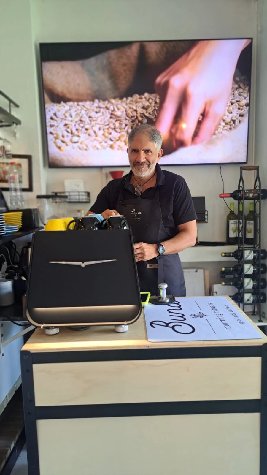 Un miembro del equipo de Buna, con delantal de la casa, sonríe tras la barra junto al tostador de café de sobremesa, con una pantalla mostrando granos de café al fondo