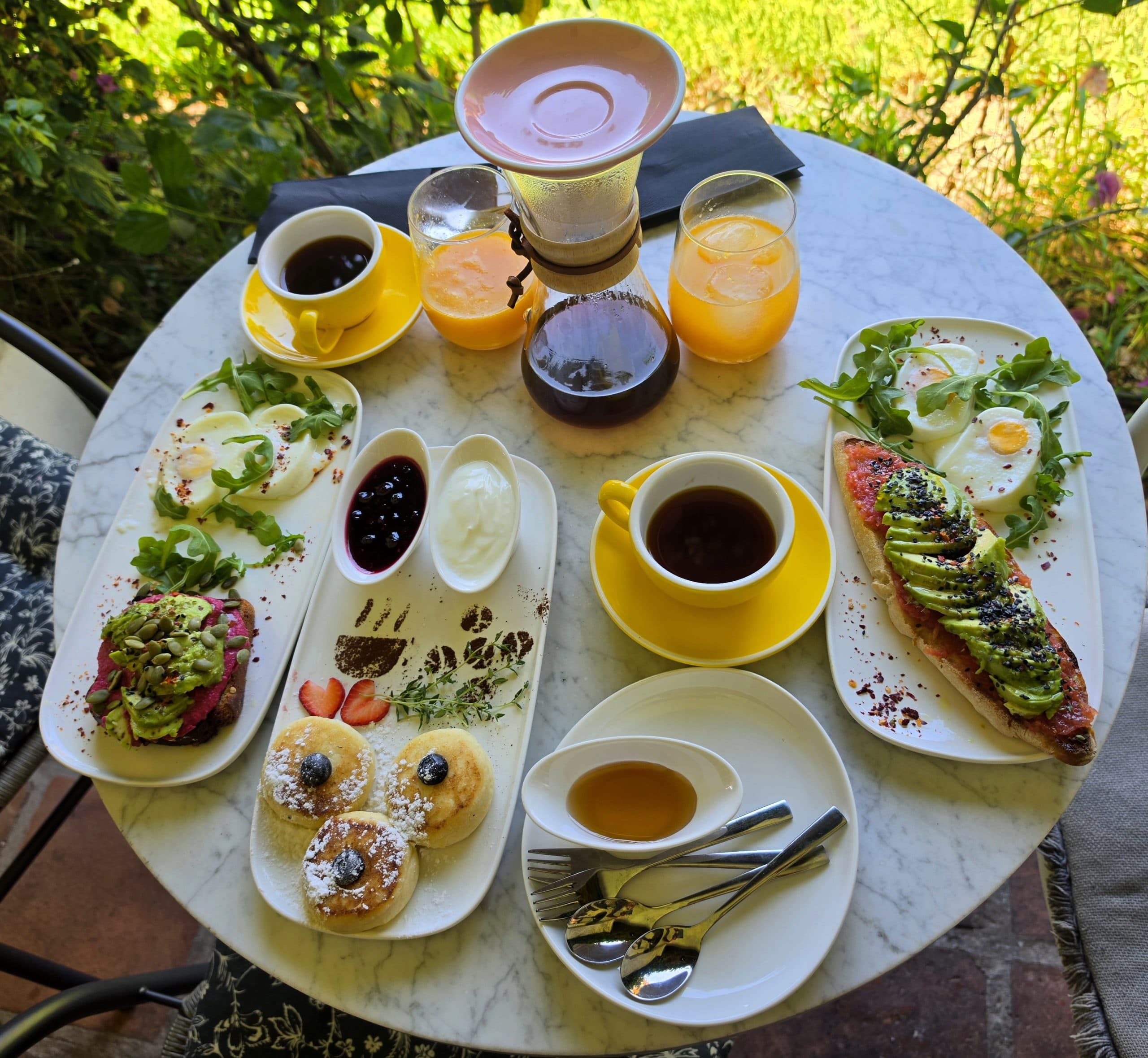 Mesa redonda de mármol con un desayuno completo: tostadas de aguacate, bollería con fruta, tazas amarillas de café y zumos de naranja