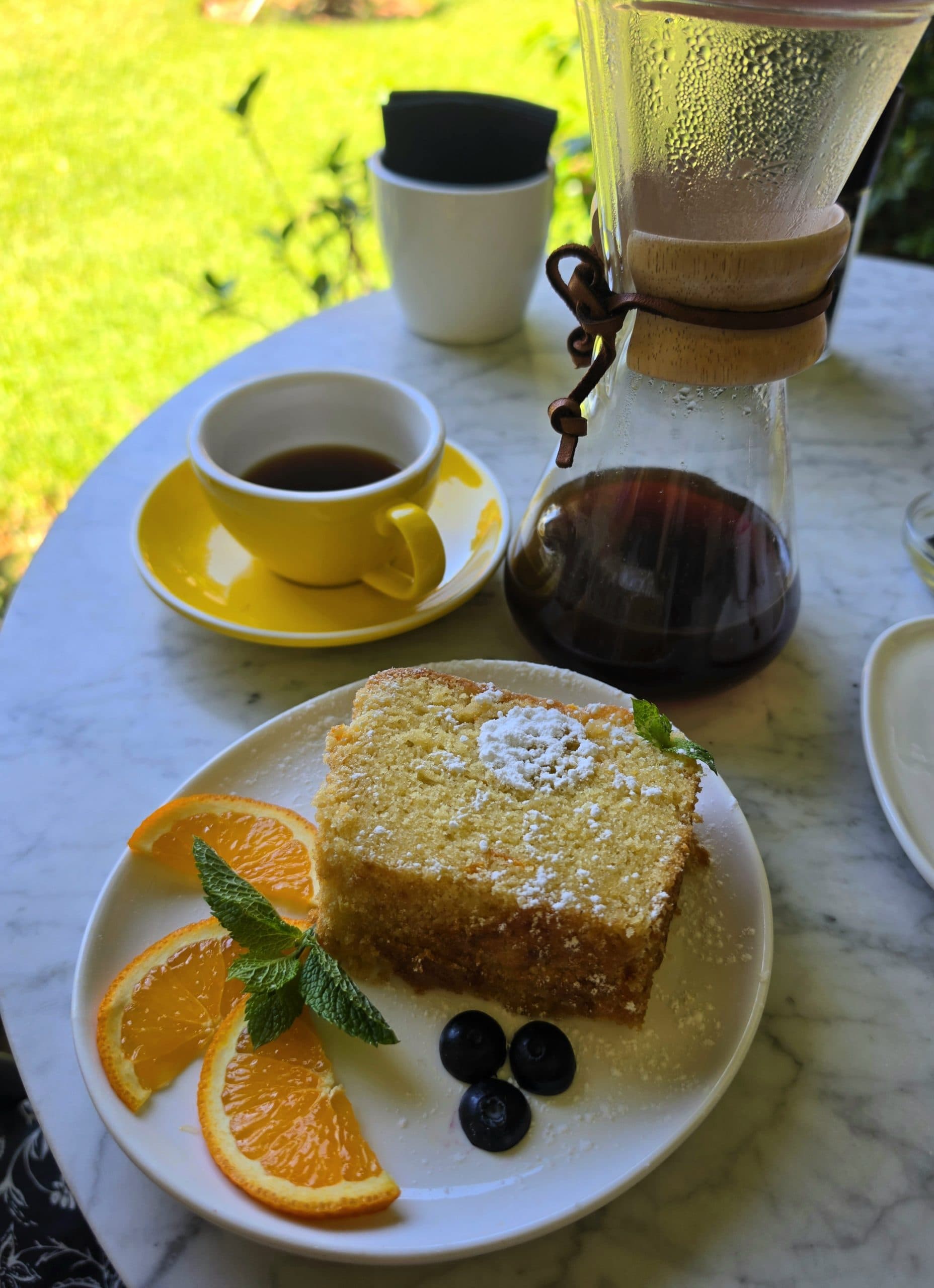 Porción de budín espolvoreada con azúcar, con rodajas de naranja y menta, junto a un café solo en taza amarilla y una Chemex, en una mesa del jardín