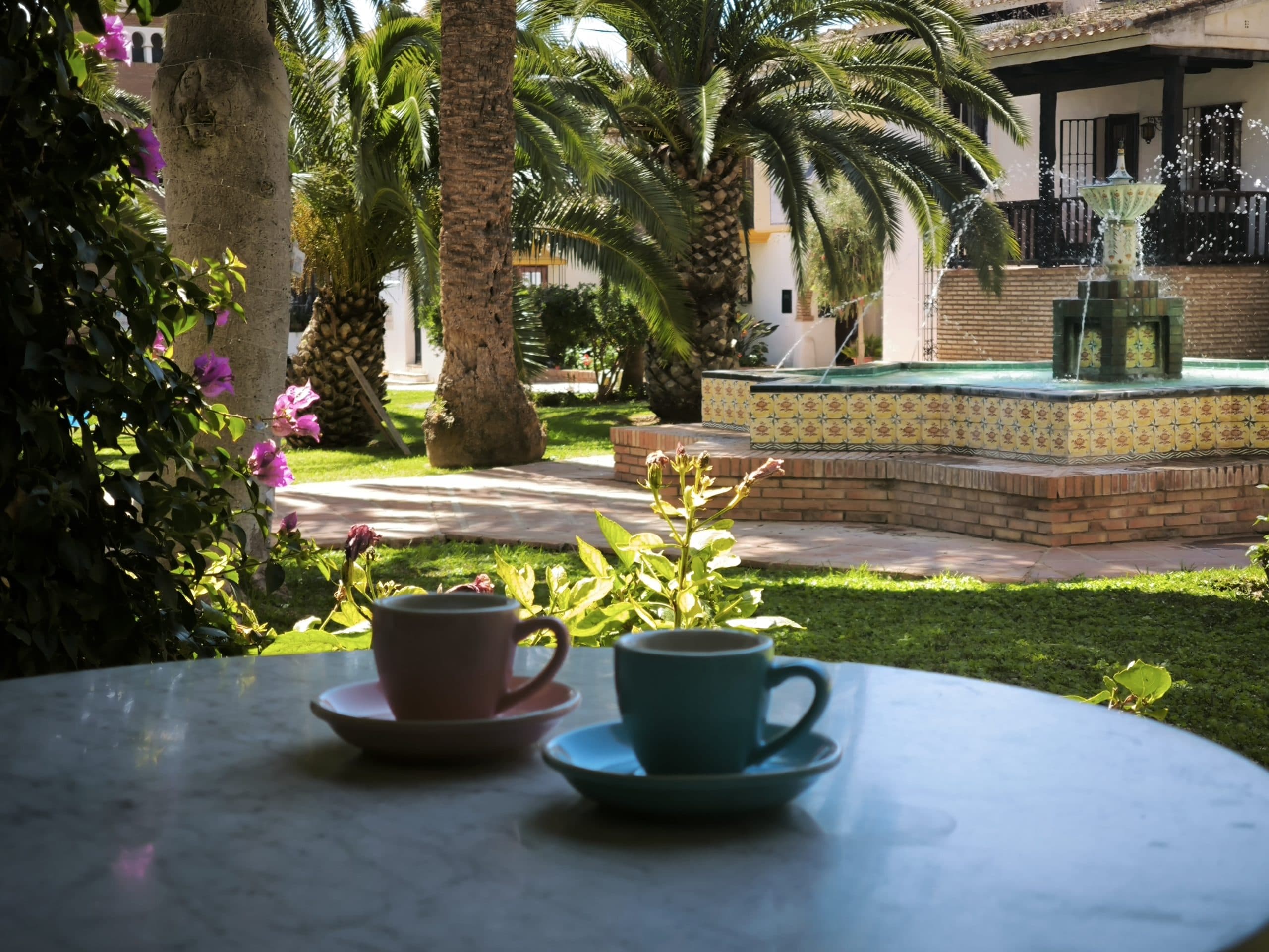 Dos tazas de café, una malva y otra turquesa, sobre una mesa redonda del jardín, con palmeras y una fuente de piedra al fondo